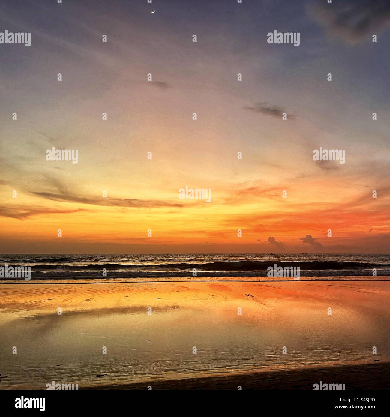 Sunset at Seminyak beach Bali with the sun set over the sea and the orange glow remaining in the clouds and the orange light reflecting in the shallow water Stock Photo