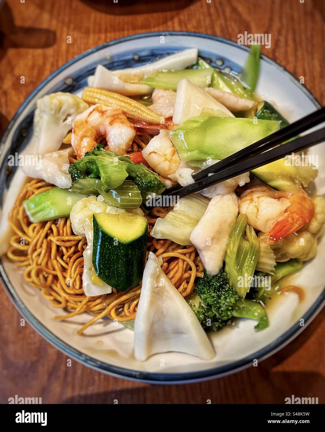 High angle close-up of stir fried noodles with seafood and mixed vegetables on plate with chopsticks on table. Chinese food. - Smartphone Captured Stock Image