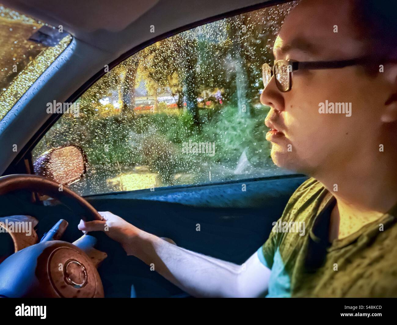 Close-up of a man at the steering wheel, driving a car on a wet, rainy night. Illumination. Street lighting. - Smartphone Captured Stock Image