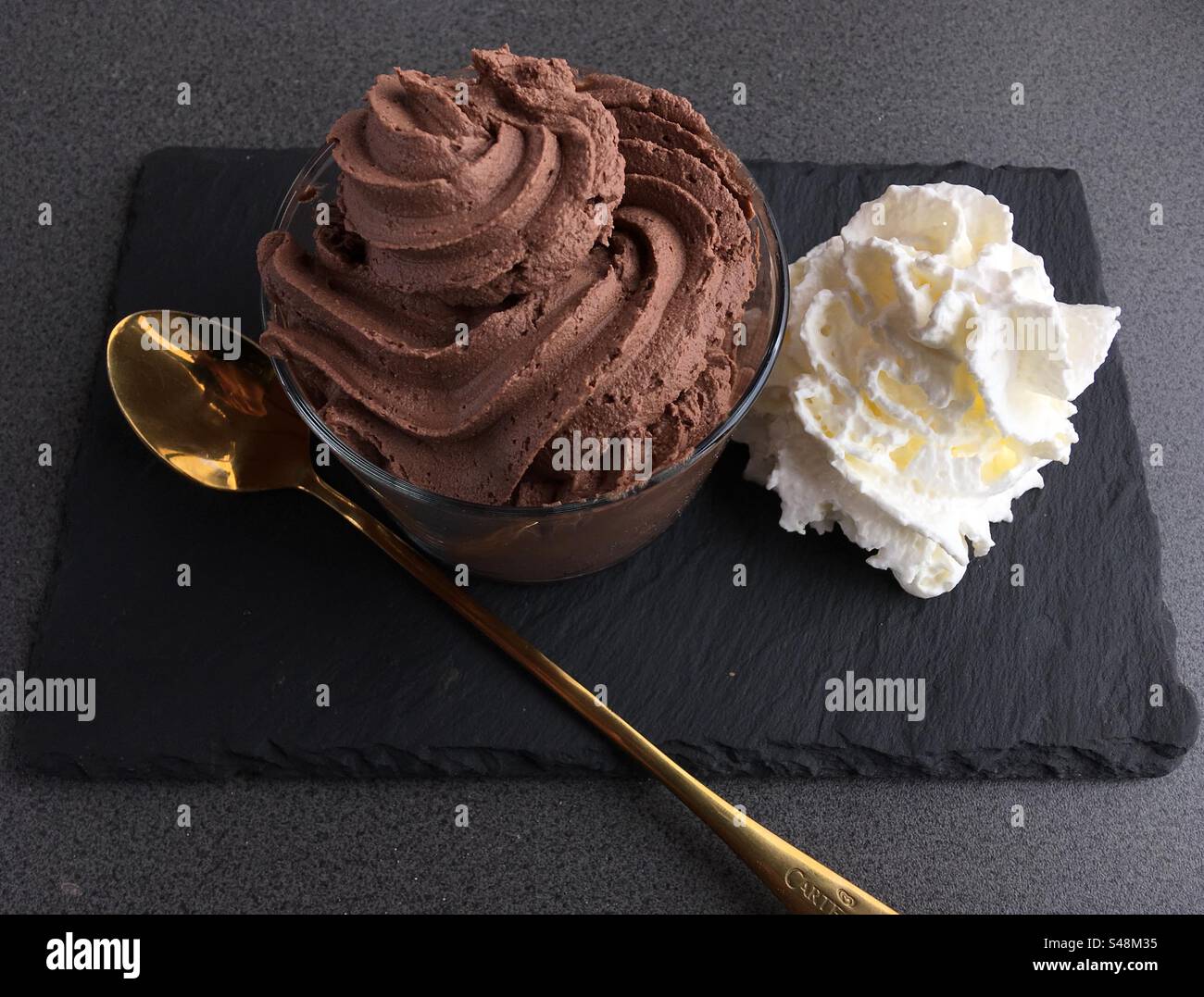 Close-up of a chocolate mousse dessert with whipped cream on a flat black stone restaurant plate with a long silver spoon - Smartphone Captured Stock Image