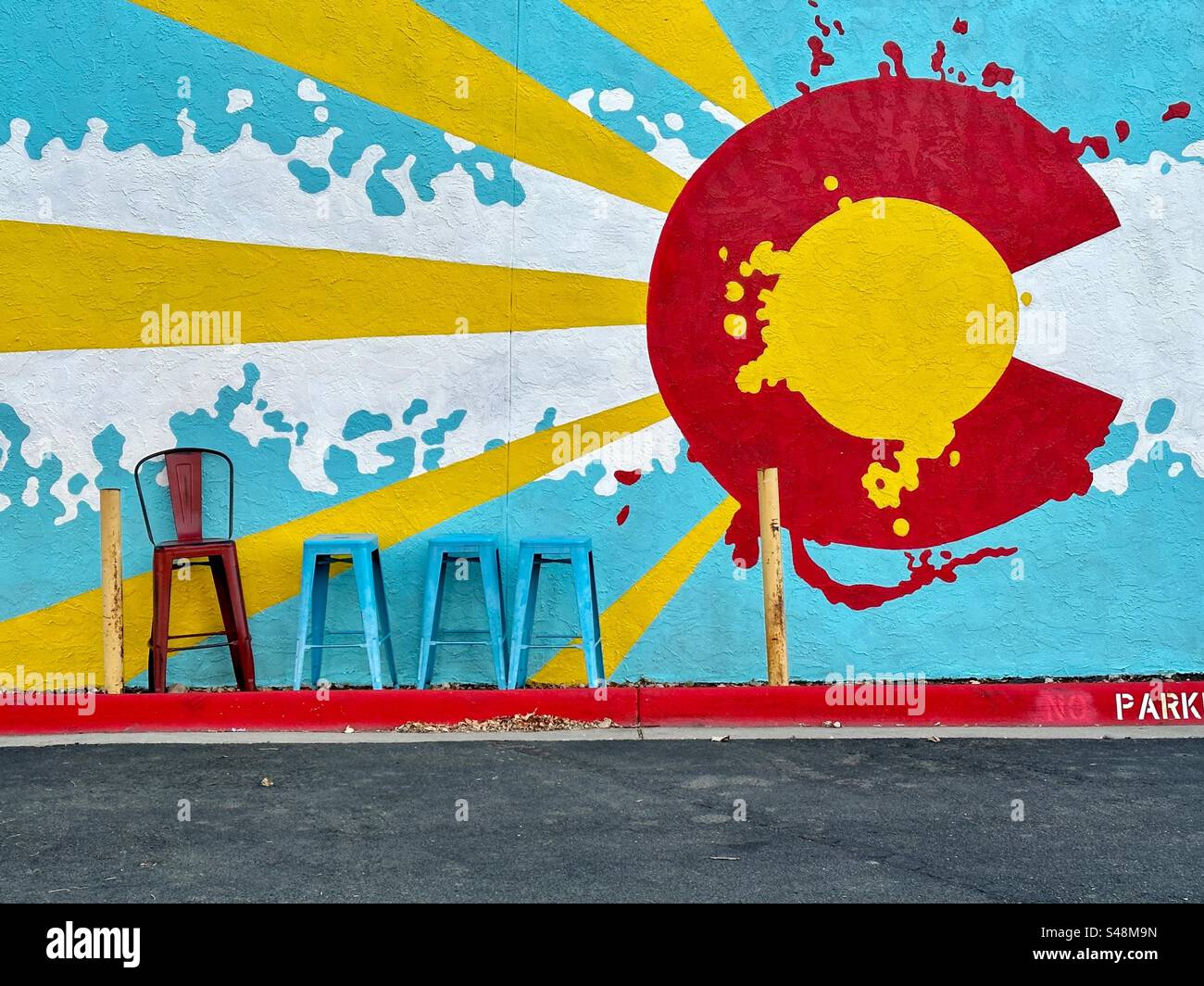 Wall mural with large red C often seen on Colorado flag. Yellow sun rays bursting out. Red chair and 3 blue stools lined up near building and parking lot. Louisville, Colorado, USA. - Smartphone Captured Stock Image