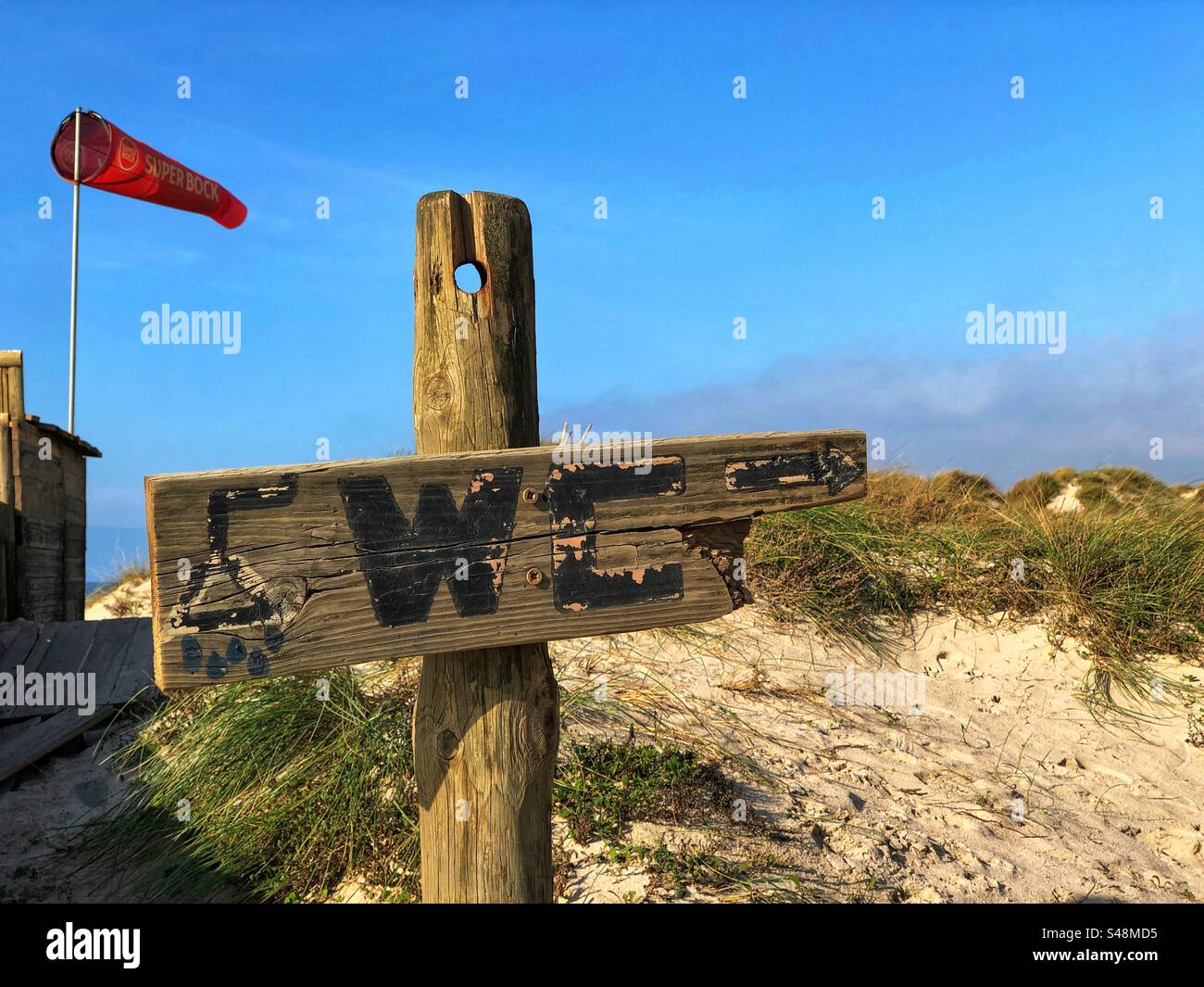 The WC wooden sign with the sand dune and the wind sock reading SUPER BOCK in the background on a sunny day in Esposendo, Portugal, November 18, 2023 - Smartphone Captured Stock Image