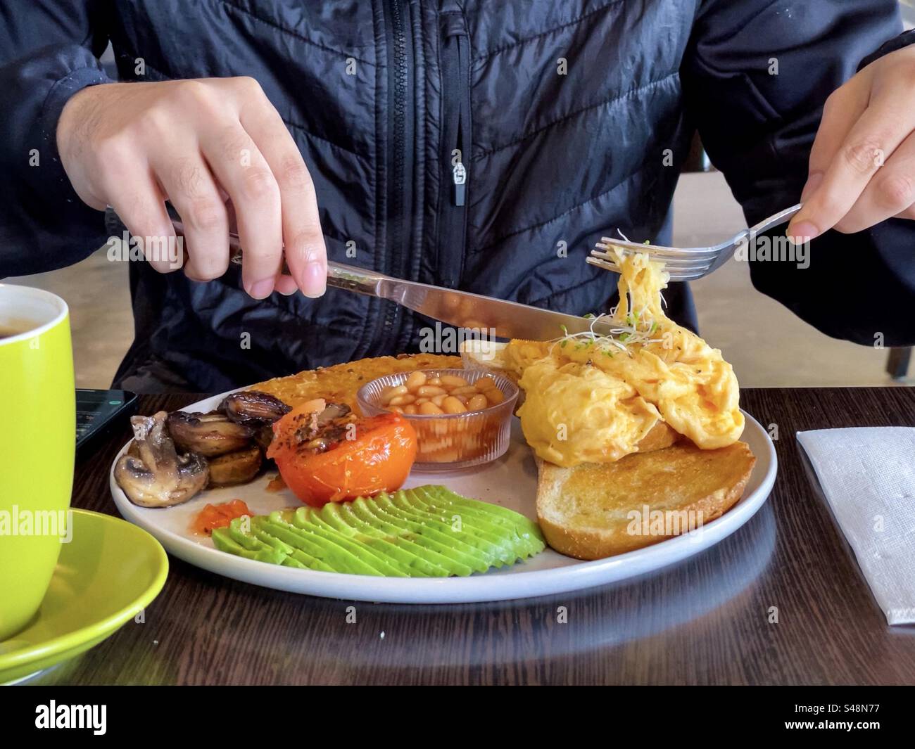 Midsection of man with hands holding cutlery and eating a healthy vegetarian breakfast of scrambled eggs on toast, grilled mushrooms and tomatoes, hash brown, baked beans and sliced avocado at table. - Smartphone Captured Stock Image
