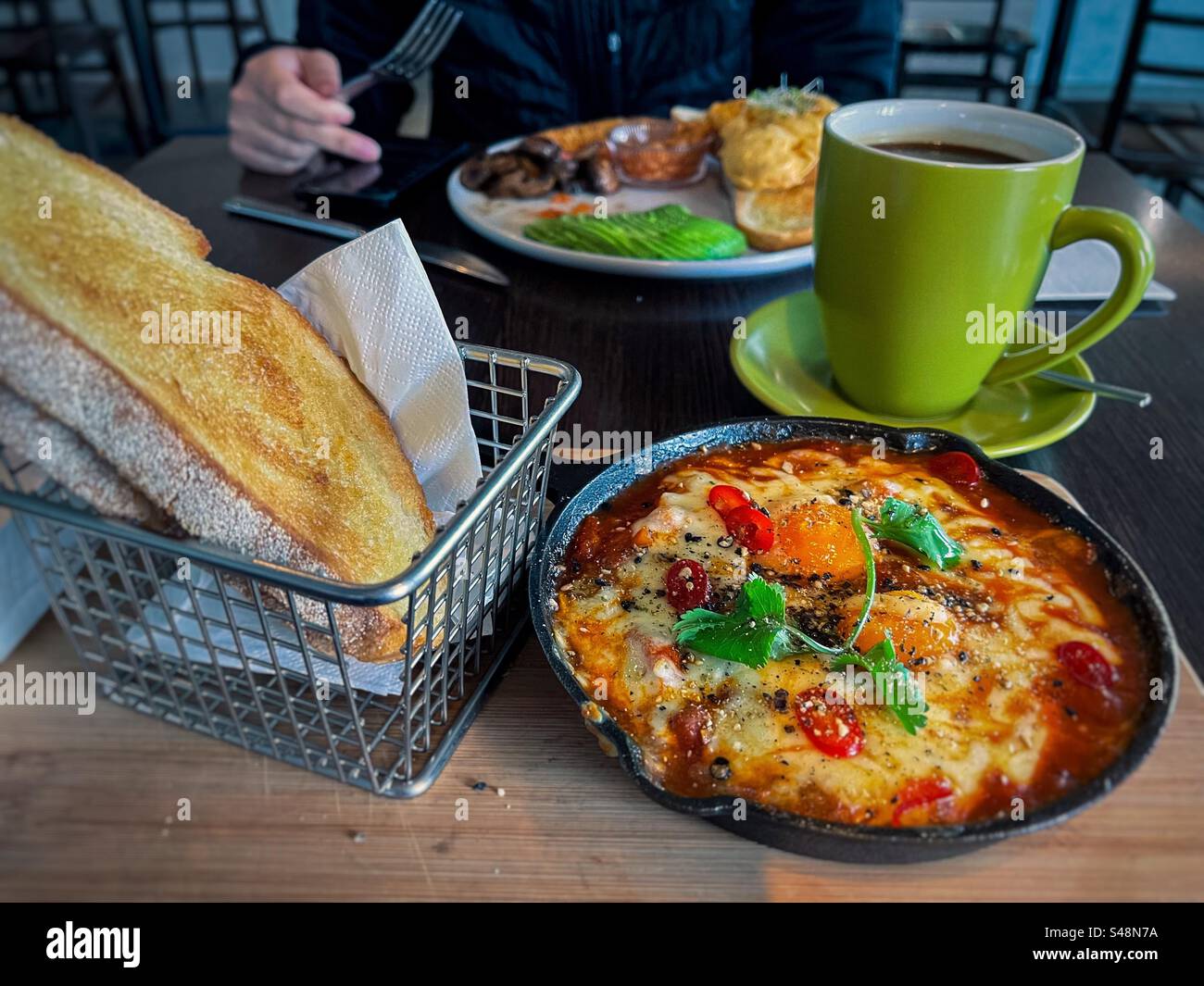 Shakshuka/poached eggs with beans in skillet with sourdough toast in basket and a cup of coffee against midsection of man holding fork and using mobile phone in a cafe. Healthy breakfast. - Smartphone Captured Stock Image