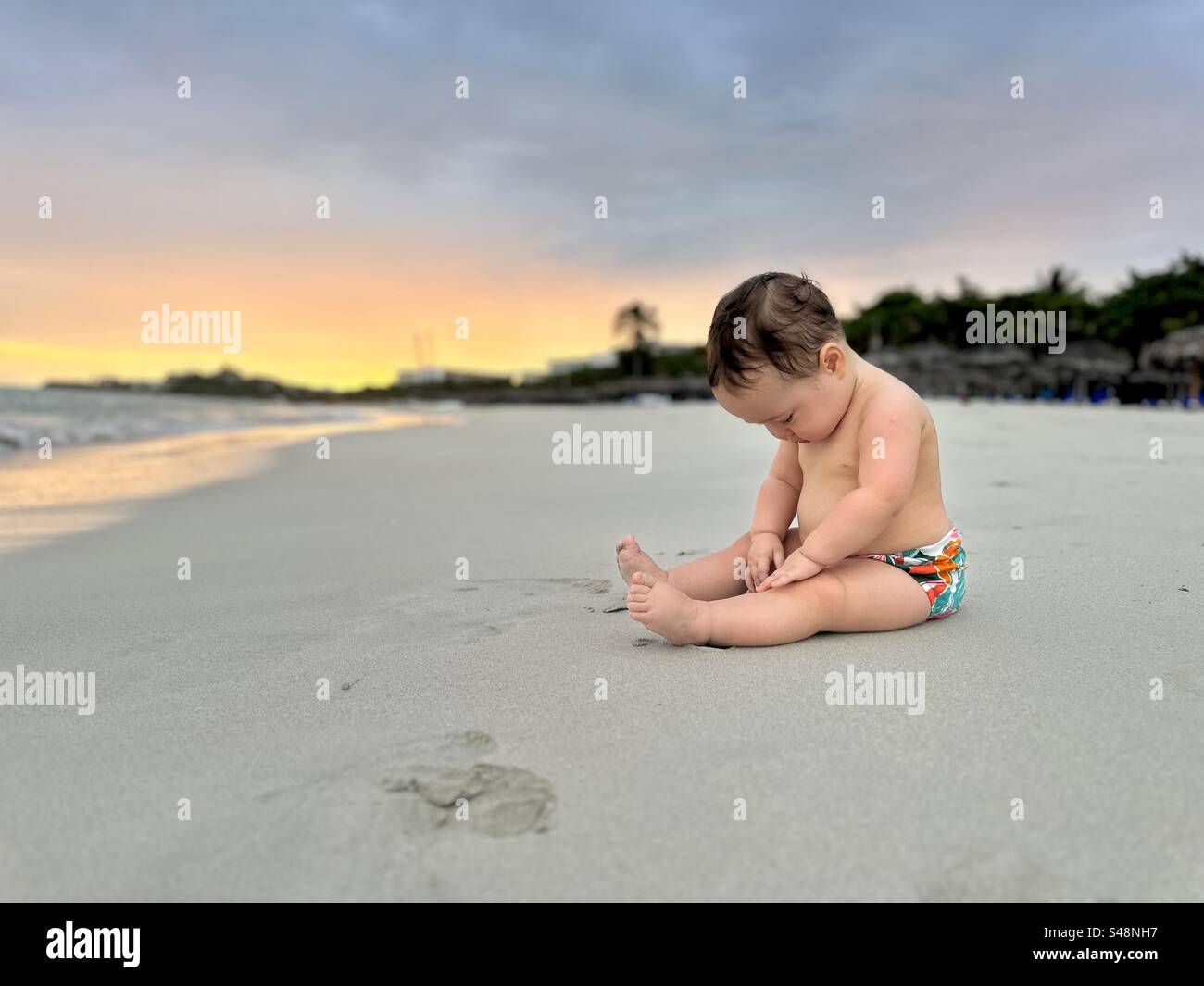Beach trip with baby. Baby sitting on the sand with face looking down. Varadero, Cuba - Smartphone Captured Stock Image
