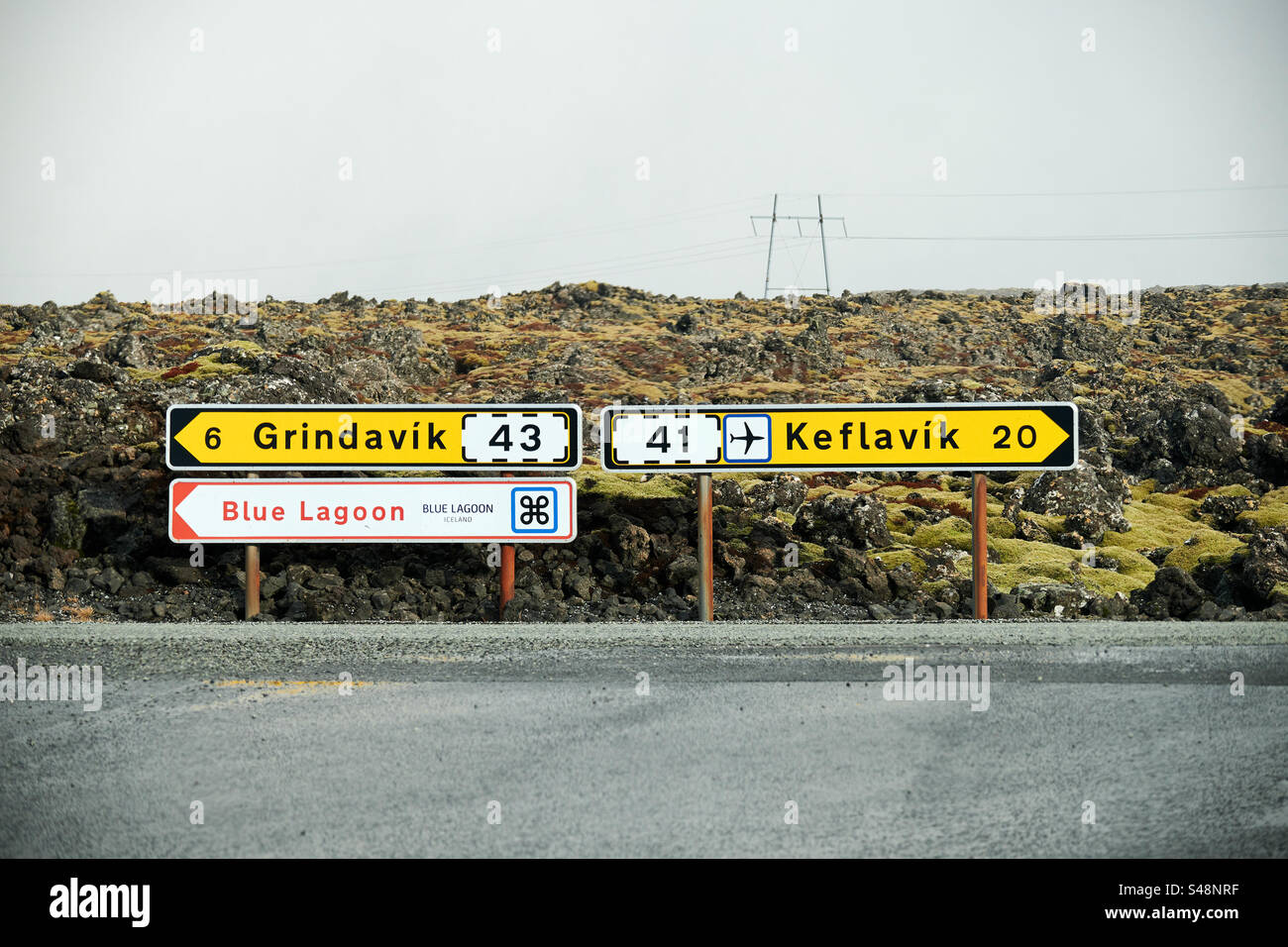 A road sign in Iceland points the way towards Grindavik, Blue lagoon, and Keflavik situated in Svartsengi. Lots of lava and moss behind it and a electric mast. Stock Photo