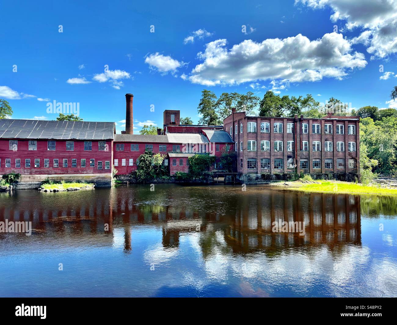 Collinsville, Connecticut, USA - July 2023:  Vintage red, manufacturing building. The former Collins Company with Farmington River in front. Reflection in water on a sunny, summer day. - Smartphone Captured Stock Image