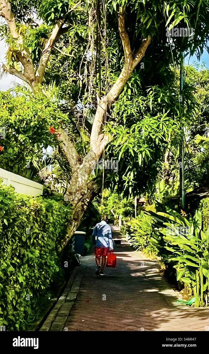Man carrying a shopping bag down a lane in Seminyak Bali with greenery of trees either side Stock Photo