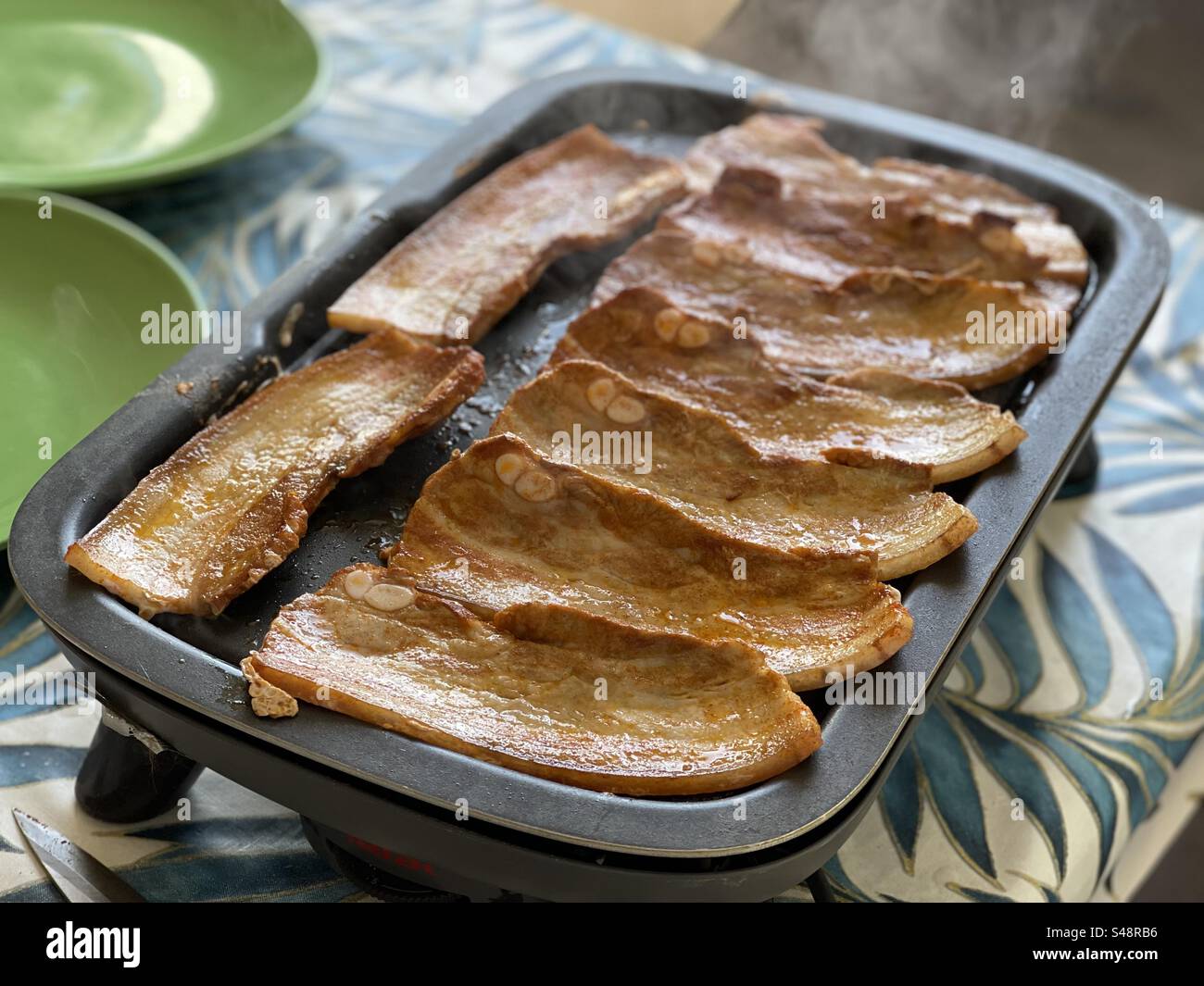 Shot of a pork belly slices on a plancha, on a blue leafy table top and green plates in France - Smartphone Captured Stock Image