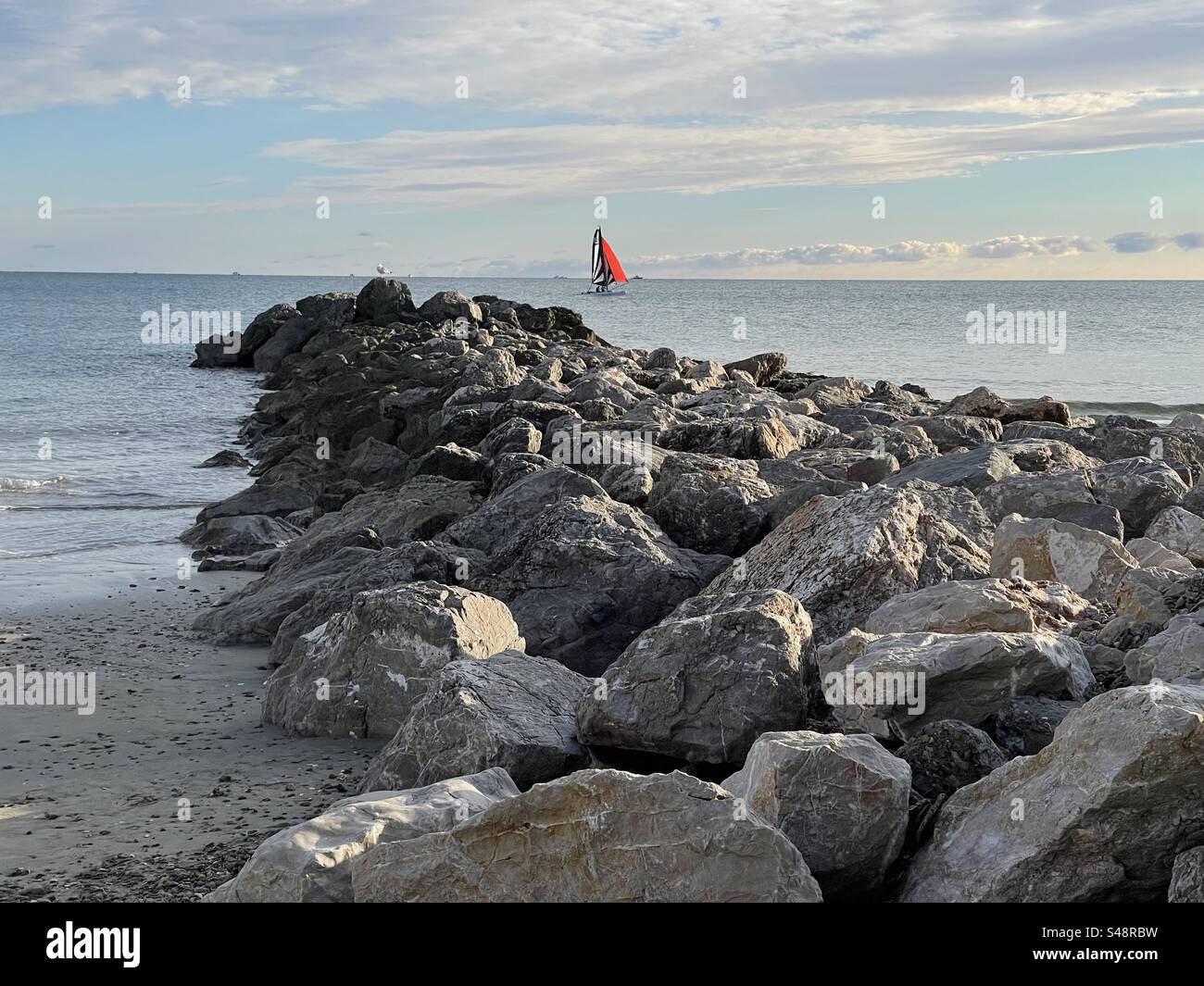 Landscape shot of a rocky pier on the Mediterranean Sea, with a red sailboat in Palavas-les-Flots, Occitanie, France - Smartphone Captured Stock Image
