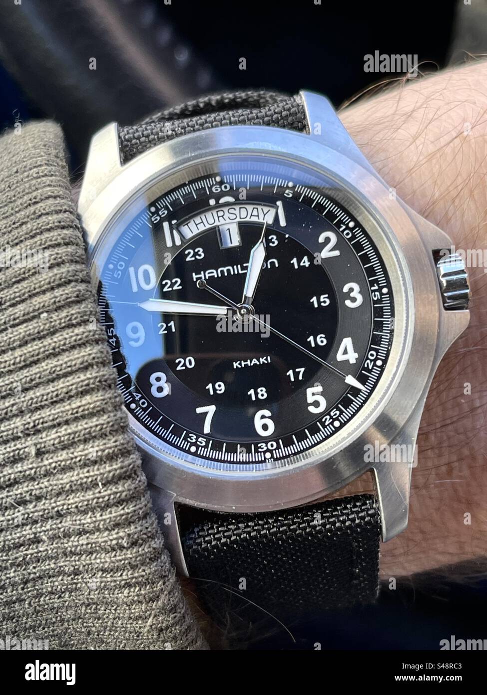 Close-up shot of a day date Hamilton Khaki military style watch with black dial on hairy wrist with a sweater on black NATO cloth strap and black background - Smartphone Captured Stock Image