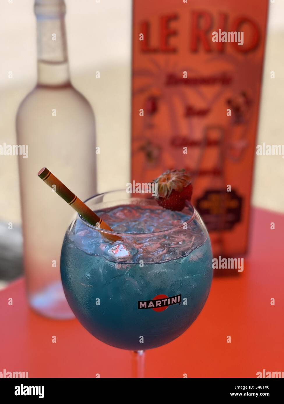 A blue lagoon cocktail in a Martini glass in front of a bottle of water and a menu for a French restaurant called Le Rio in Palavas-les-Flots, Occitanie, France Stock Photo