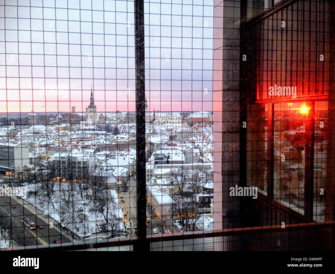 Beautiful view through the window with metal mesh lattice over the roofs of the winter Tallinn city with the red sunrise reflecting in the next window - Smartphone Captured Stock Image