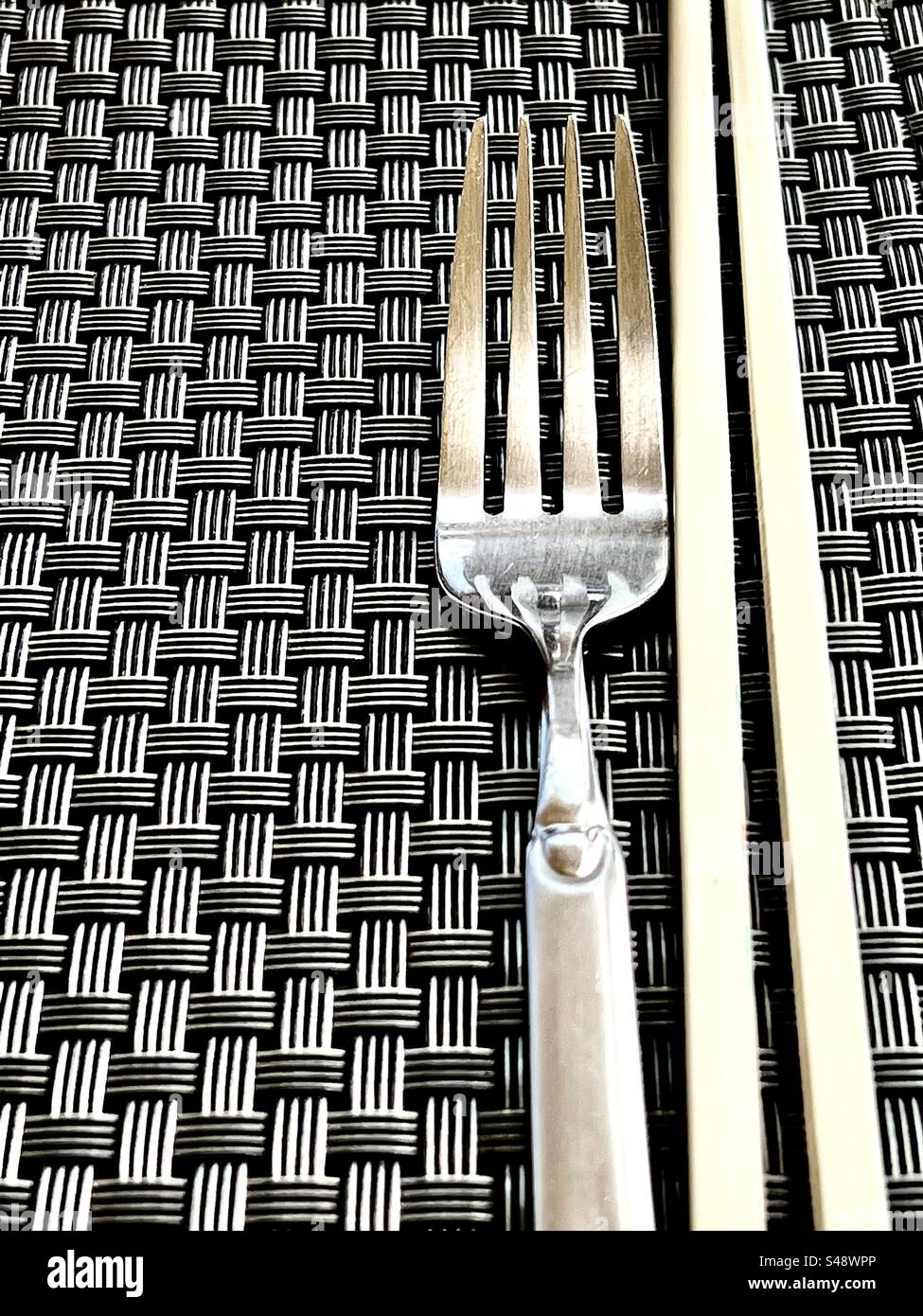 Sharp, high contrast photo of a fork and two chopsticks on a criss-cross square pattern table mat. - Smartphone Captured Stock Image