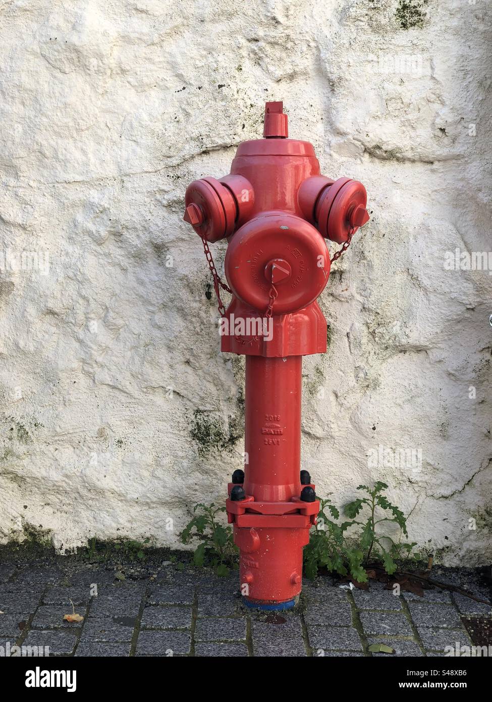 Viana do Castelo, Portugal - December 01, 2023: A vibrant red fire hydrant with chains stands against a textured white wall on a cobblestone street - Smartphone Captured Stock Image