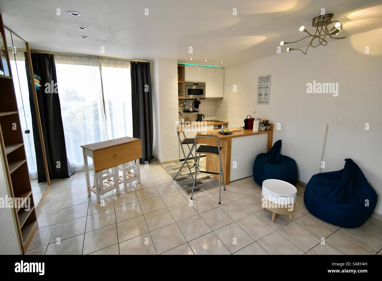 Interior shot of a bright Airbnb studio apartment with table and chairs, open concept American kitchen and white floor tiles. - Smartphone Captured Stock Image