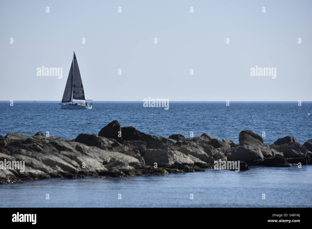 Sailboat sailing away beyond the Rocky pier on the Mediterranean Sea in Palavas, Occitanie, France. - Smartphone Captured Stock Image