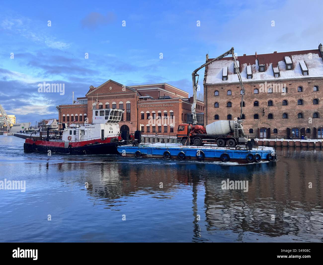 Tugboat pushing a floating platform pontoon with a concrete truck on Motlawa River by the Polish Baltic Philharmonic in the Old Town of Gdansk, Poland - Smartphone Captured Stock Image