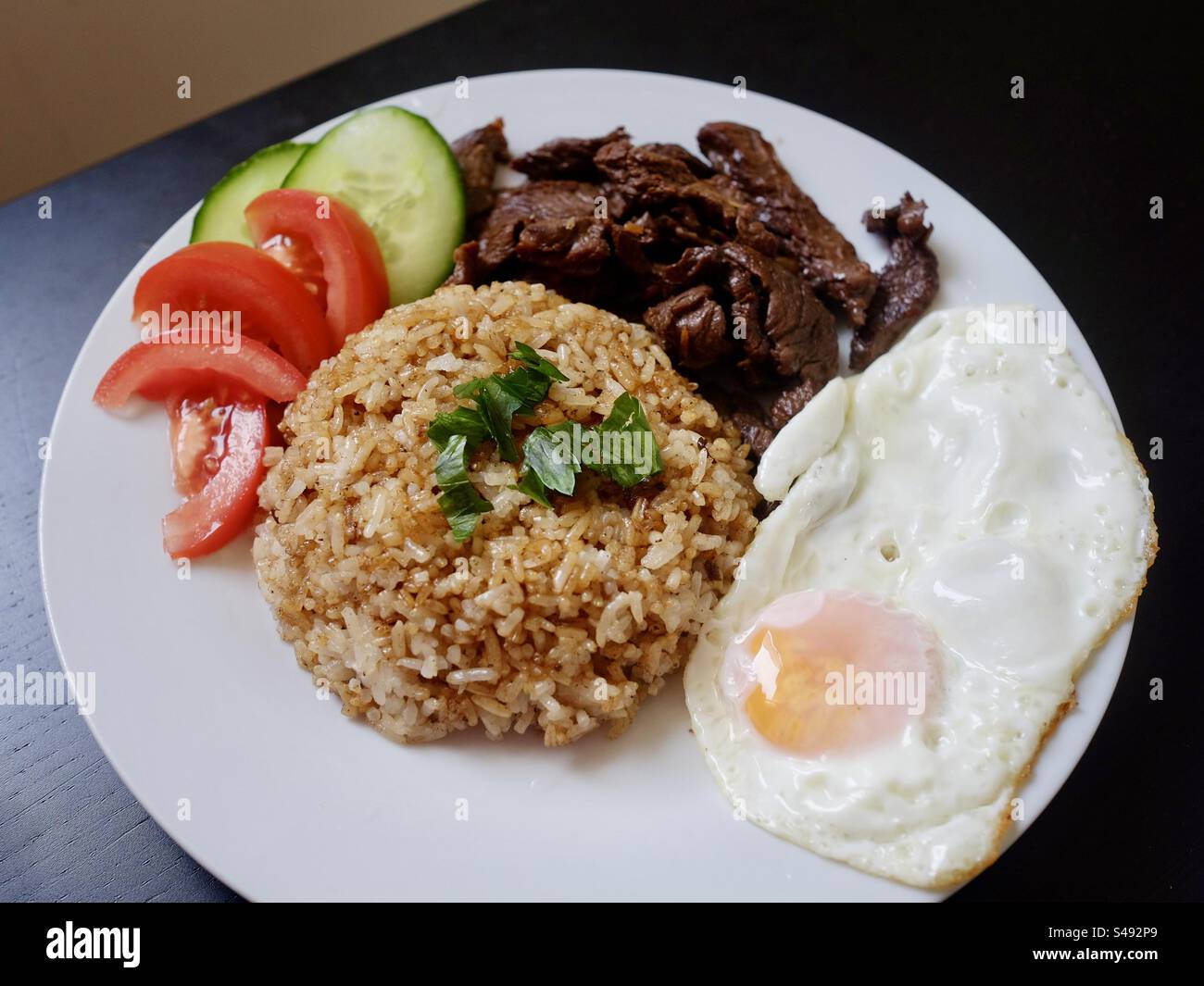 A white porcelain plate with homemade fried rice, egg sunny side up, sliced barbecue beef asian style, cucumber and tomato slices on a black wooden table backdrop. - Smartphone Captured Stock Image
