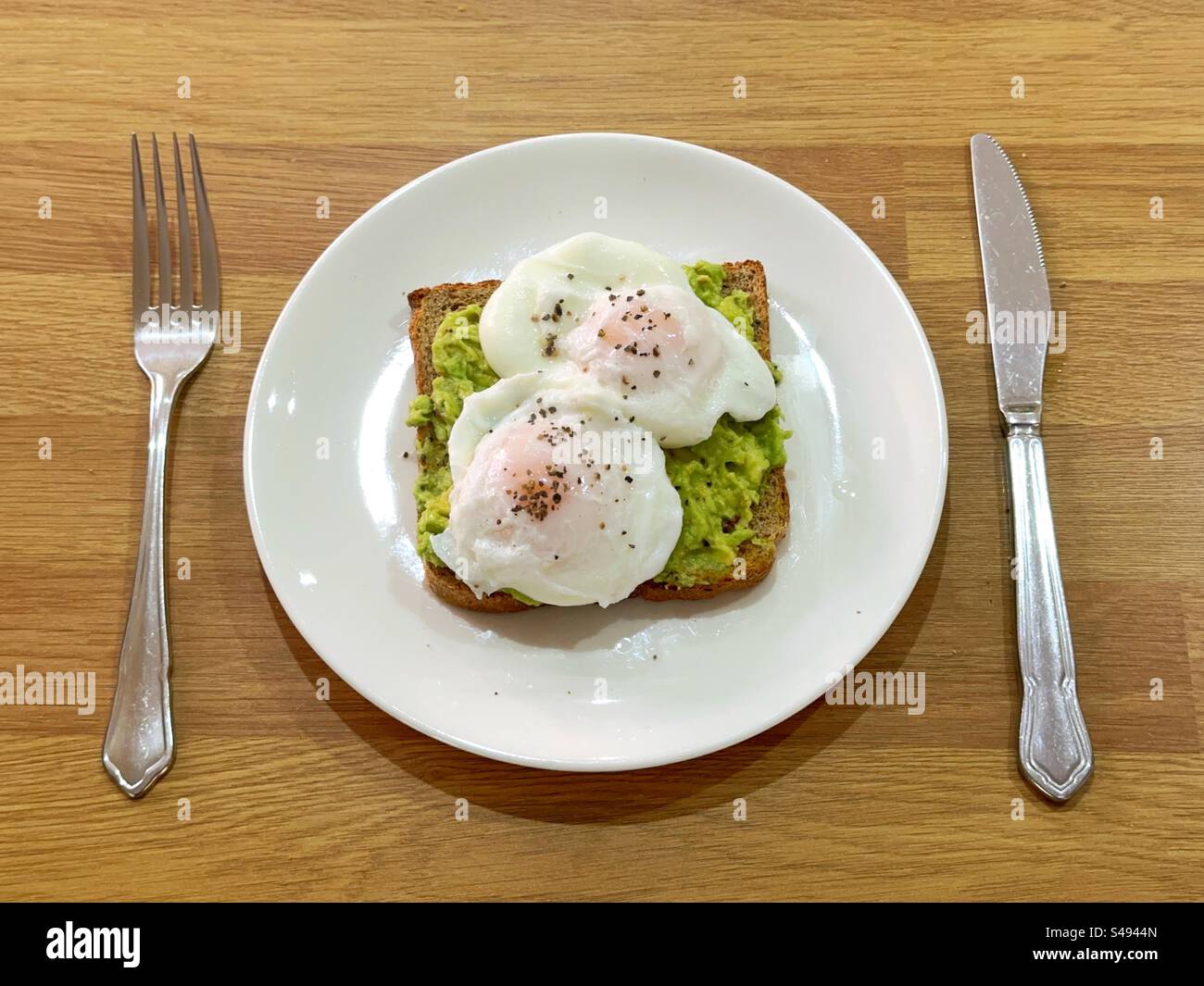 Poached eggs on avocado toast sprinkled with black pepper on a white plate between a knife and fork. - Smartphone Captured Stock Image