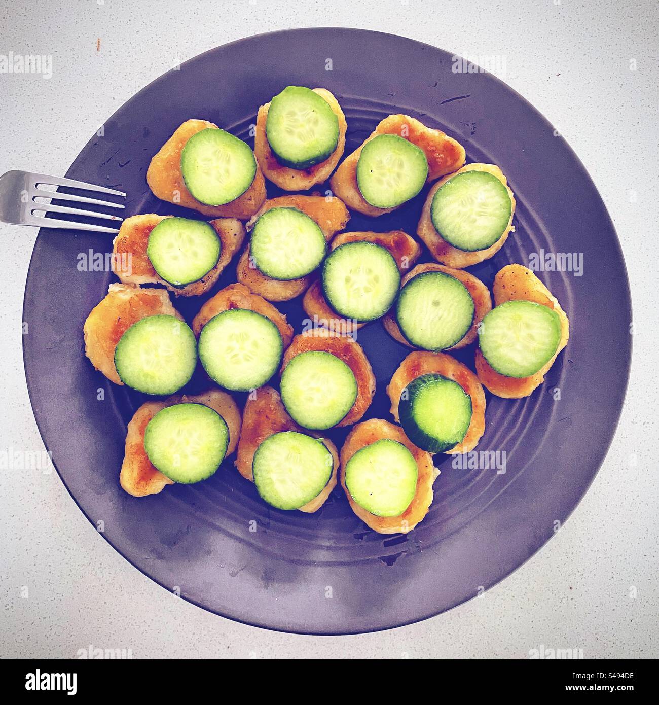 Top view of plate of many fried chicken nuggets with slicse of green cucumber on each with afork nearby teady to be eaten for lunch diner by hungry starving kid child children food - Smartphone Captured Stock Image