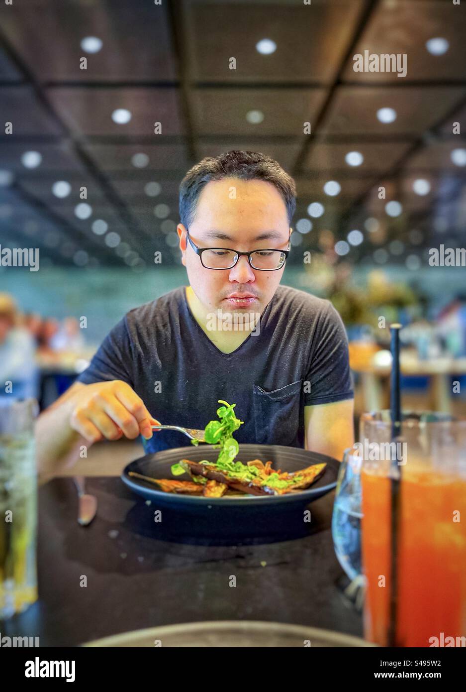 Asian man in eyeglasses enjoying a vegetarian meal of fresh salad greens and miso roasted eggplants at table in restaurant. Healthy eating. Focus on foreground. Stock Photo