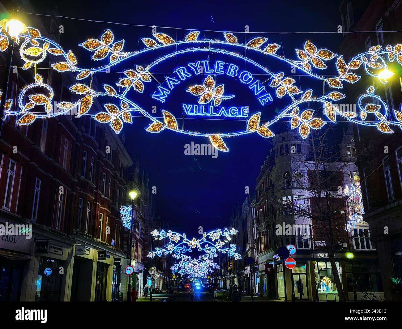 Christmas lights across the street read: “Marylebone Village” the relative low rise building of historic London at night. - Smartphone Captured Stock Image