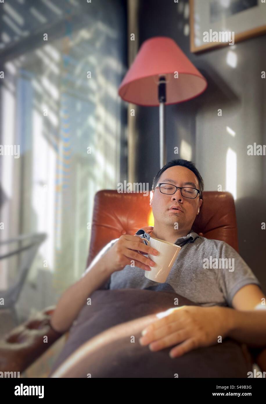 Asian man with wireless earbuds, listening to music and relaxing in armchair with a coffee cup against wall and glass door. - Smartphone Captured Stock Image