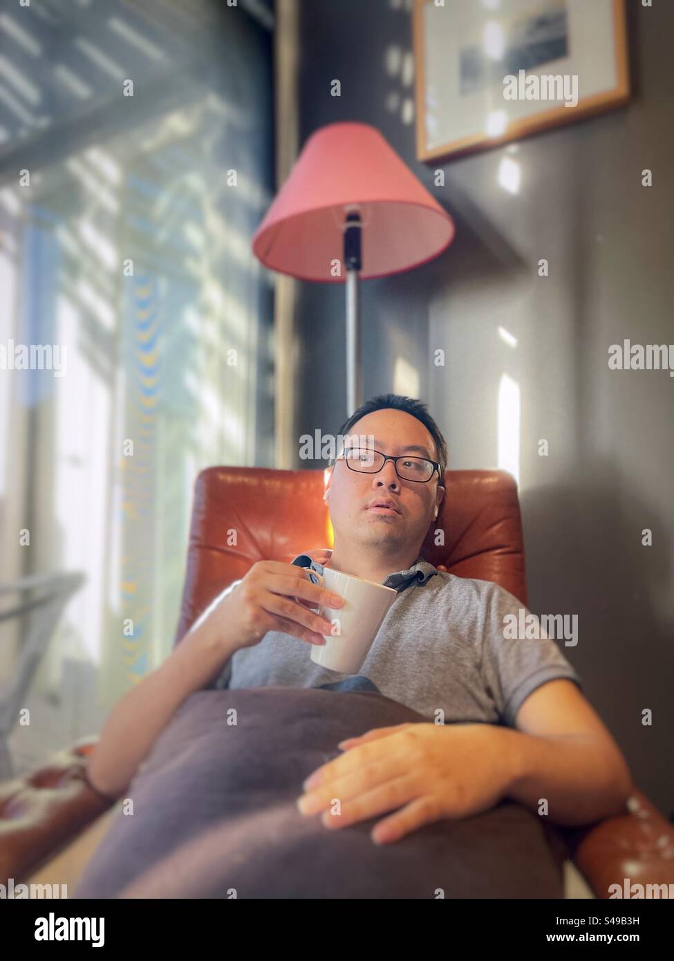 Asian man with wireless earbuds, listening to music and relaxing in armchair with a coffee cup against wall and glass door. - Smartphone Captured Stock Image