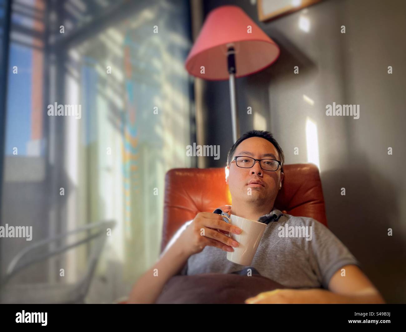 Asian man with wireless earbuds, relaxing in armchair with a coffee cup against wall and glass door. - Smartphone Captured Stock Image