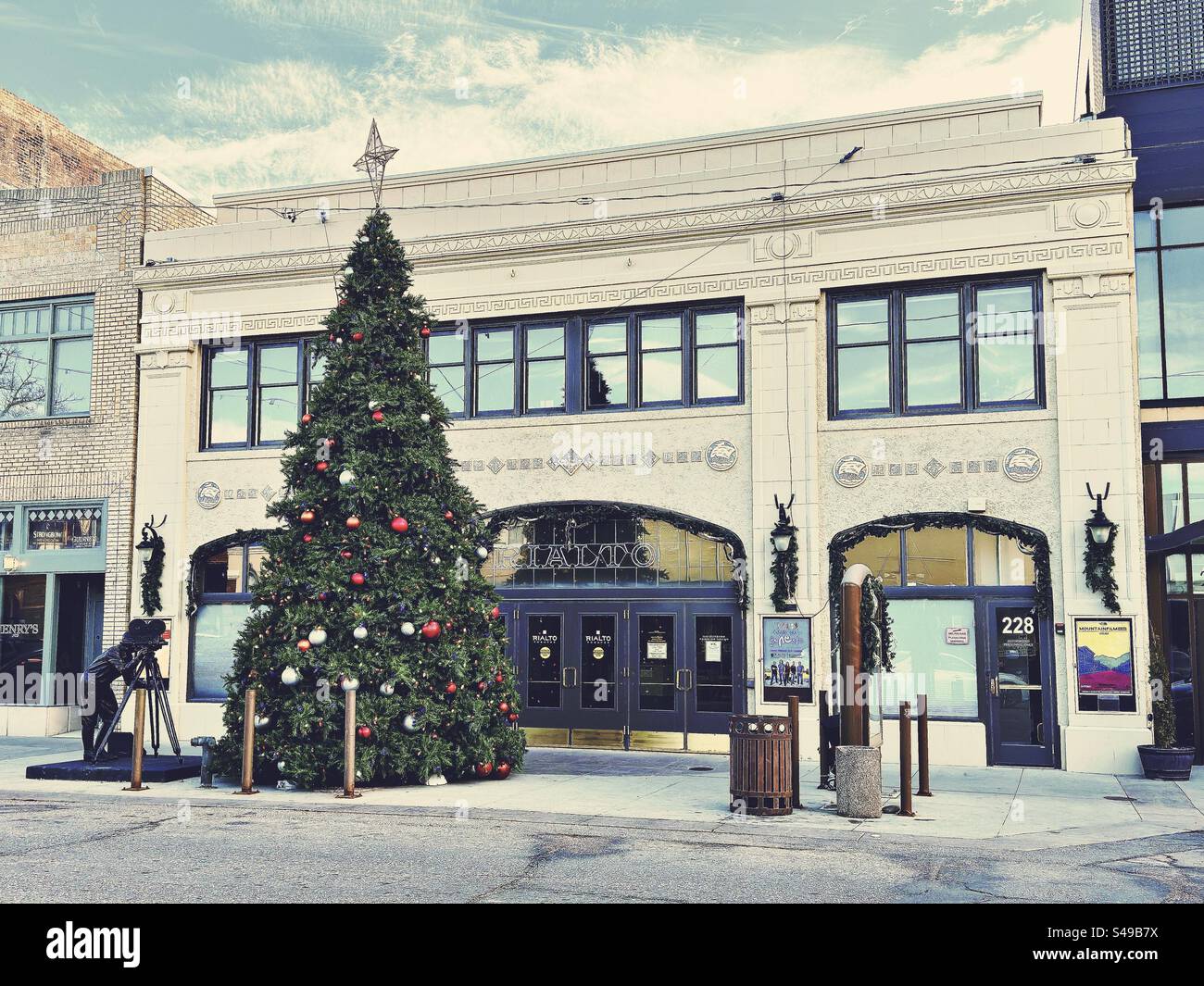 Tall Christmas tree in downtown Loveland, Colorado, USA during winter. Vintage architecture with street view. Statue of man with movie camera next to holiday, evergreen decoration. - Smartphone Captured Stock Image