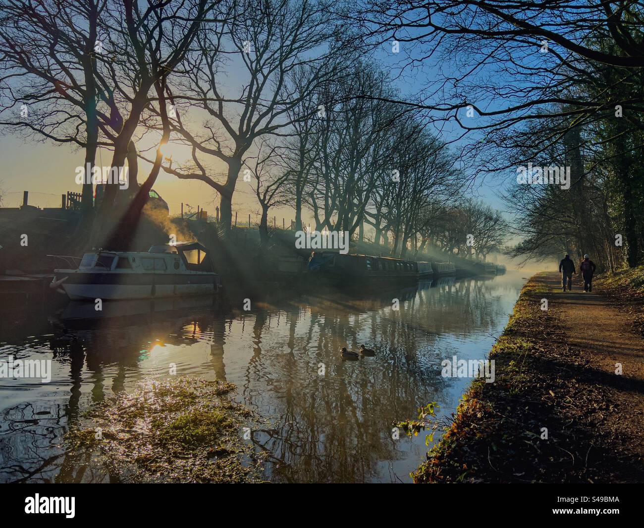 Reflections in winter morning mist on Leeds and Liverpool canal at Adlington, Lancashire. Two people walking on tow path with reflections of trees and narrow boats in canal. Stock Photo