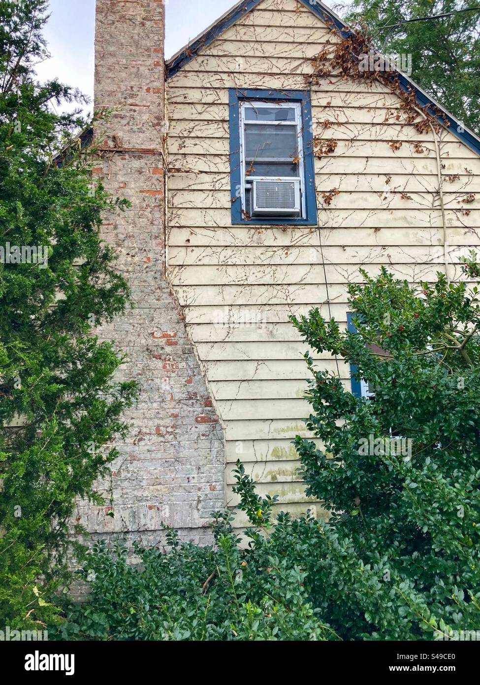 Exterior of old, wooden home in Madison, Connecticut, USA. Second story window has air-conditioning unit. Chimney is attached to outside of building and paint is worn out. Trees around the house. - Smartphone Captured Stock Image
