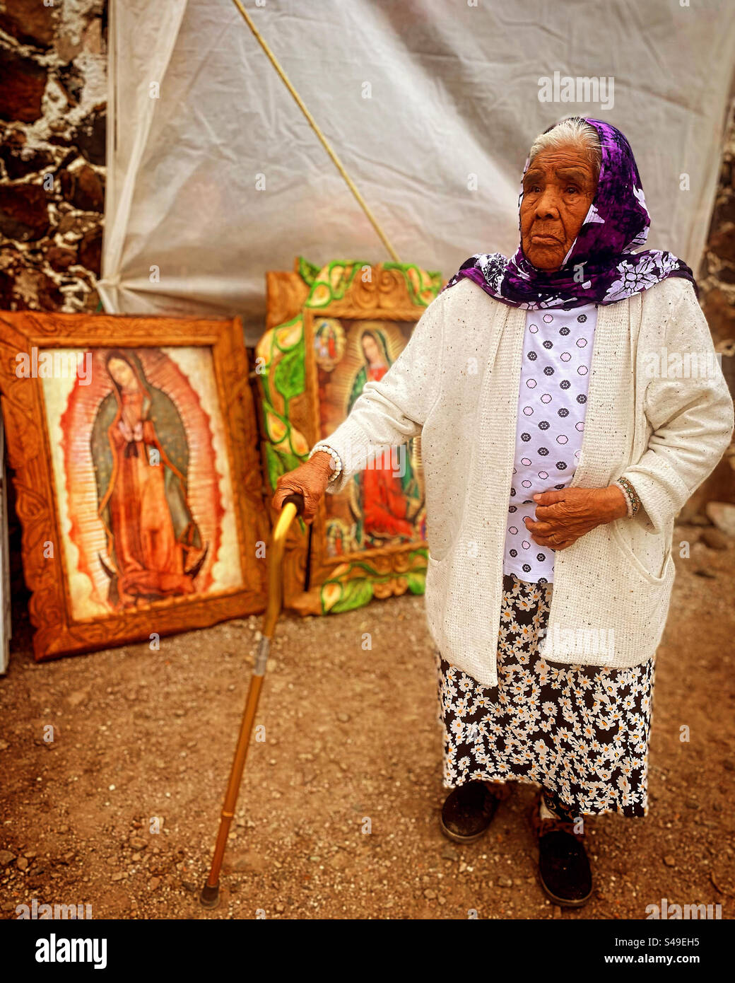 A Mexican elderly woman using a walking stick and a head scarf stands in front of images of Our Lady of Guadalupe in Colon, Queretaro, Mexico - Smartphone Captured Stock Image