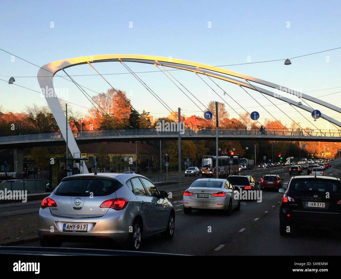 Perspective view to the stylish modern design footbridge over the road in the low autumn sun light from the street with several cars, directions signs, lamps on the wires and the tram behind - Smartphone Captured Stock Image Perspective view to the stylish modern design footbridge over the road in the low autumn sun light from the street with several cars, directions signs, lamps on the wires and the tram behind - Smartphone Captured Stock Image