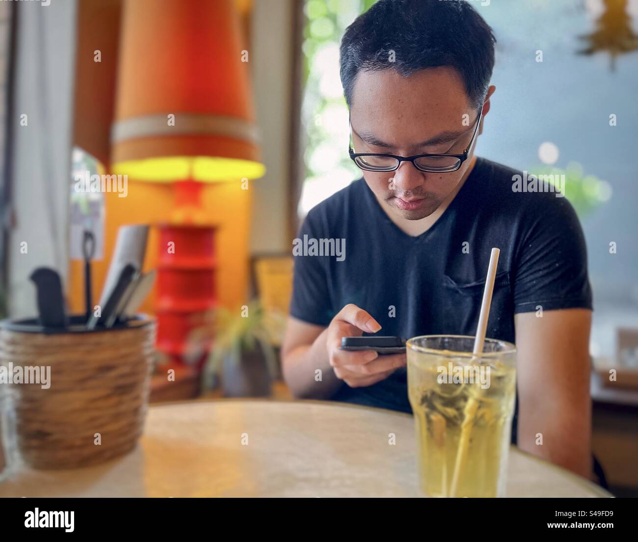 Asian man in eyeglasses using a mobile phone while having a soft drink at table in cafe. Selective focus. - Smartphone Captured Stock Image