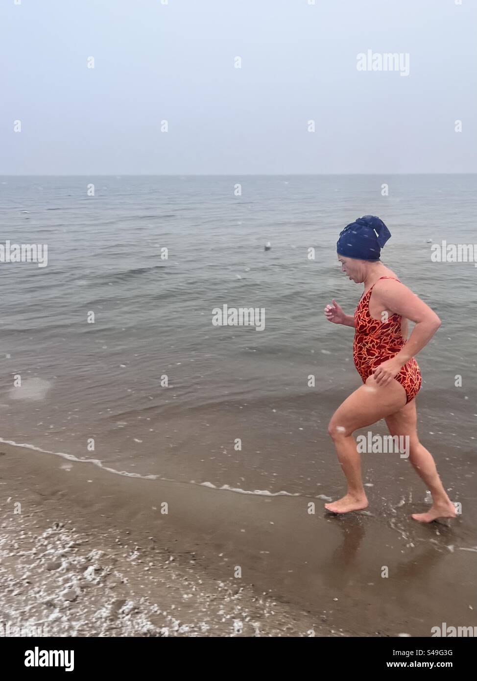 Lone female Winter swimmer in bright swimsuit jogging alongside the seashore at the Baltic Sea Gdansk beach during a snowfall in Gdansk, Poland, Europe, EU Stock Photo