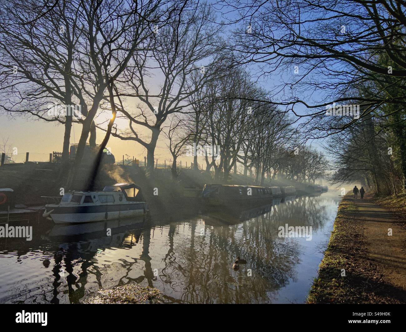 Misty winter morning on Leeds and Liverpool canal at Adlington in Lancashire. Narrow boats moored by bank and two people walking on tow path with sun peeping from behind tree. - Smartphone Captured Stock Image