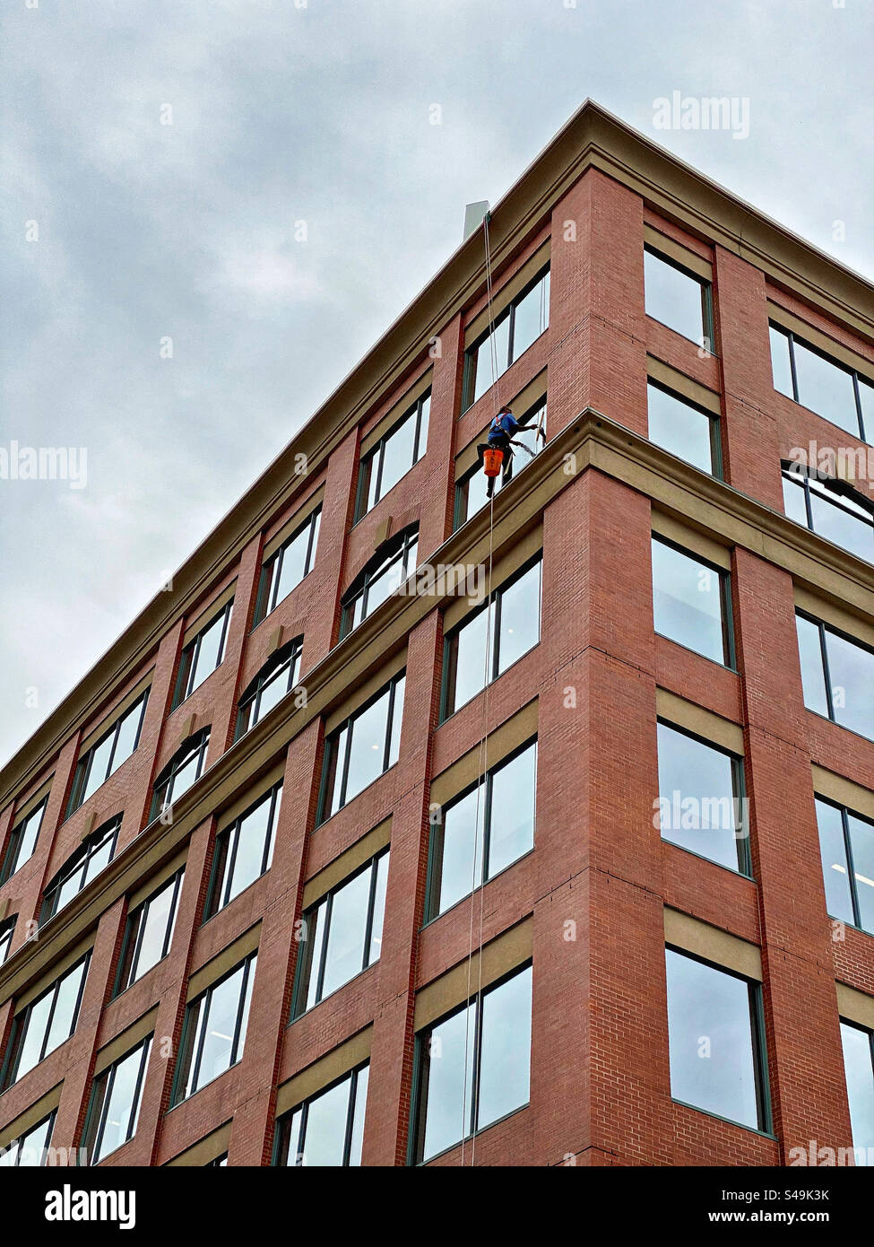 A window washer works alone, suspended from the roof of a tall brick building. Stock Photo