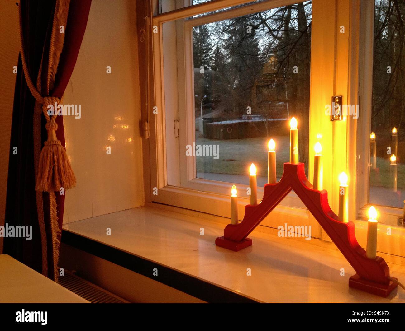 Cozy, tranquil and warm indoor interior view through the window with curtain and red triangle candlestick on the wide white windowsill to the outside twilight - Smartphone Captured Stock Image