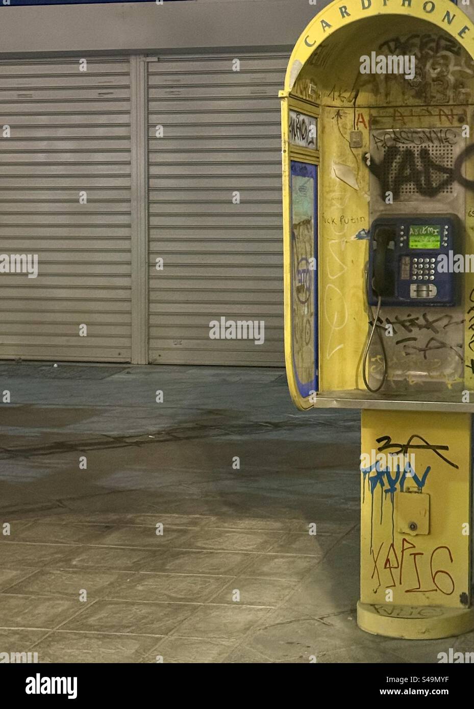 Graffitied yellow payphone booth in an urban street at night, symbolizing urban decay and outdated technology on street in the Athens historic center - Smartphone Captured Stock Image