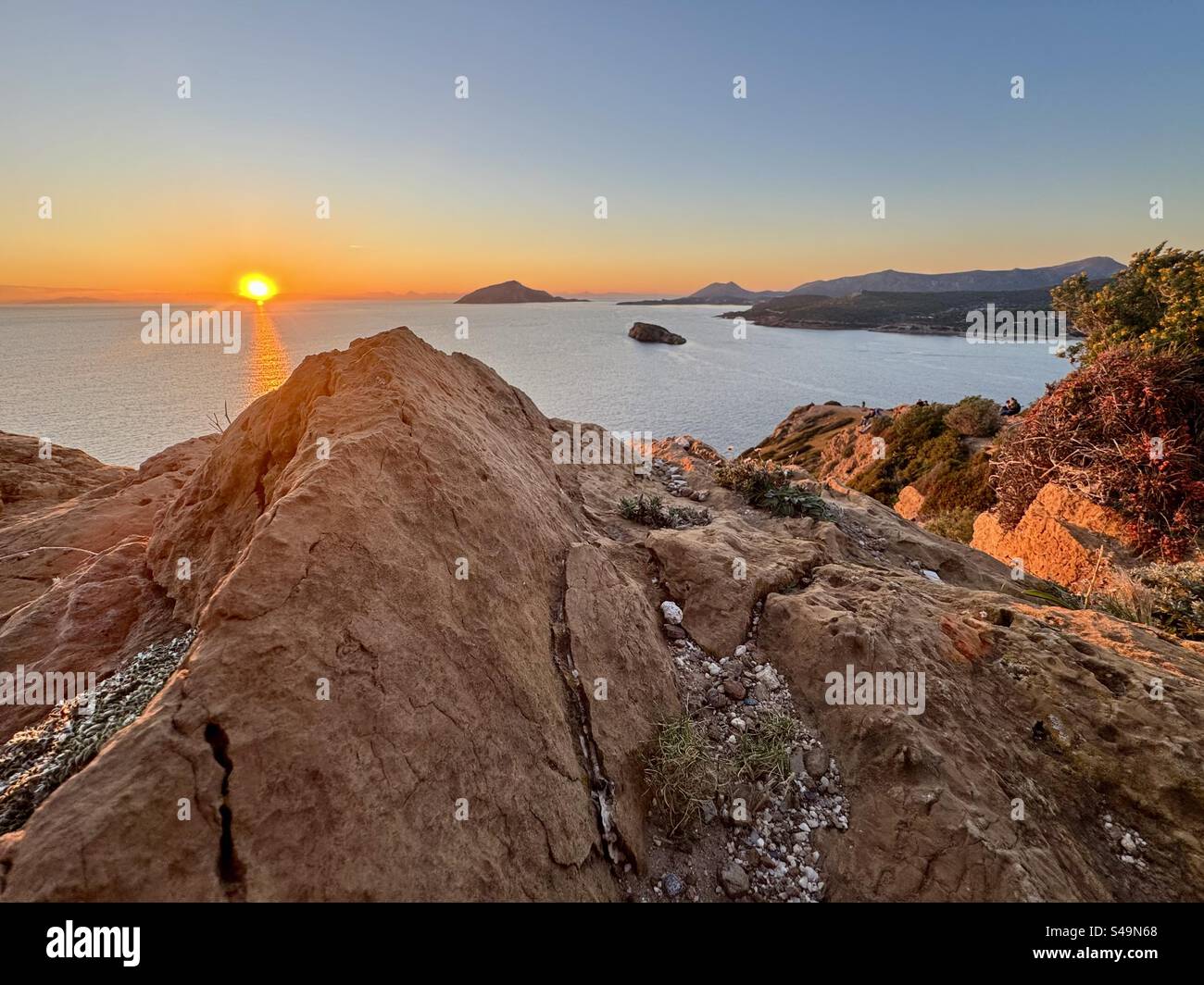 A scenic view from Cape Sounion, showcasing a golden sunset over the Aegean Sea from the rocky cliffs, with distant islands and coastline in view. - Smartphone Captured Stock Image