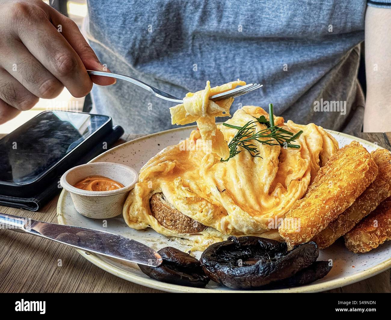 Midsection of man eating breakfast of scrambled eggs on sourdough toast, hash browns and grilled mushrooms on plate on table. - Smartphone Captured Stock Image
