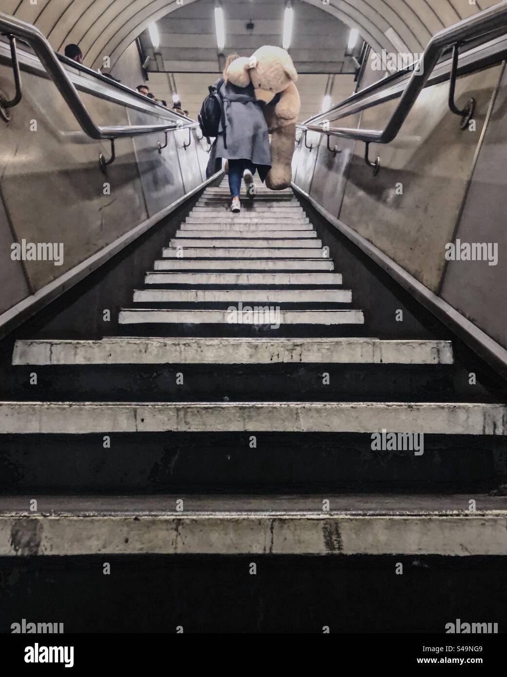 Woman walking up London Underground stairs with giant Teddy Bear over shoulder - Smartphone Captured Stock Image