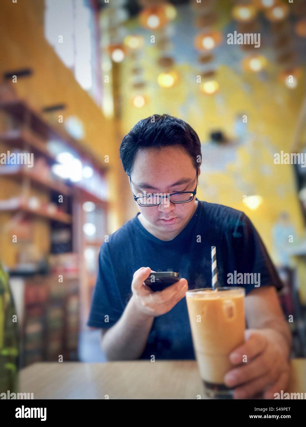 Asian man in eyeglasses using mobile phone while drinking a glass of iced tea at table in cafe. Wireless technology. Food and drink. - Smartphone Captured Stock Image