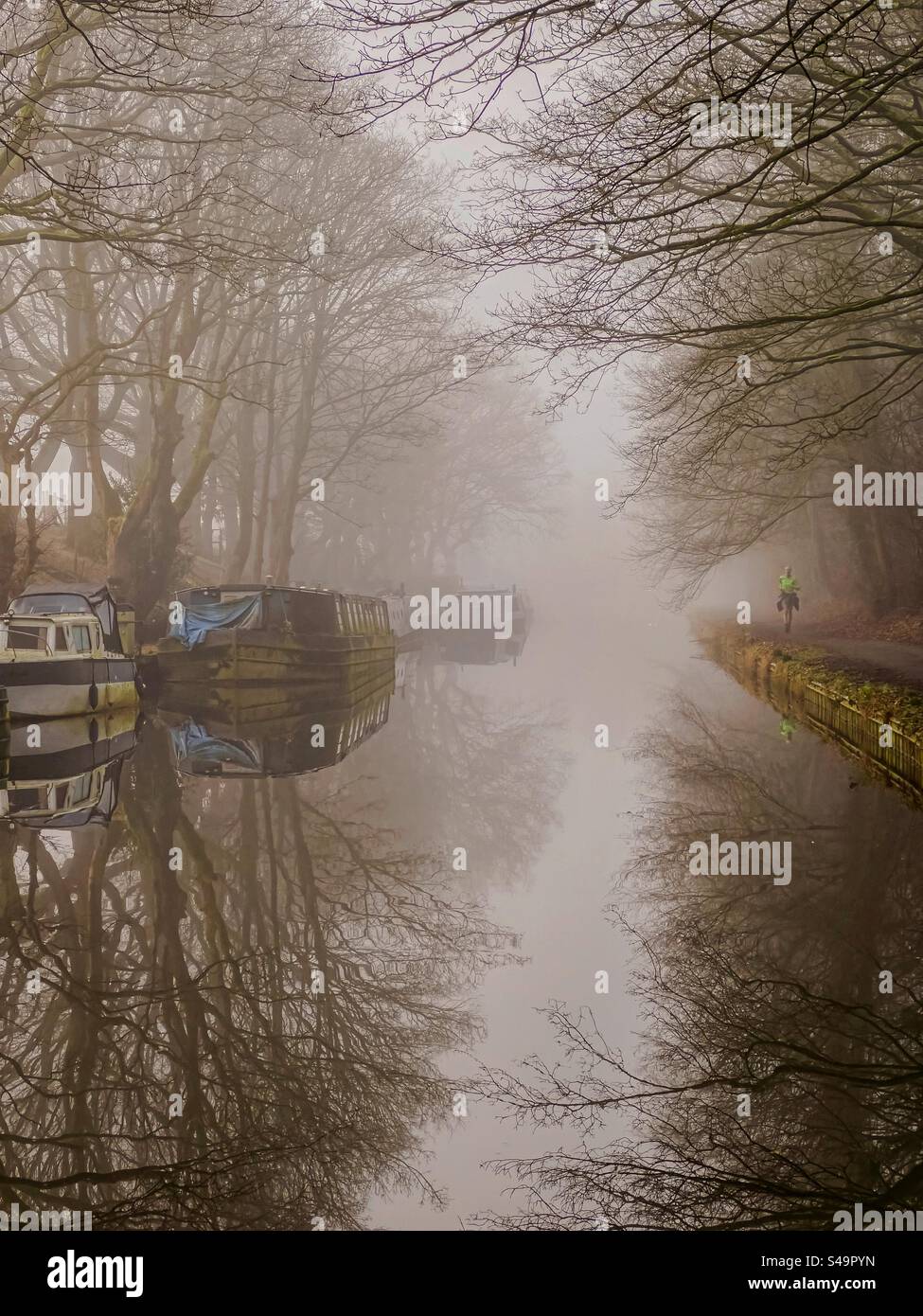 Runner on the tow path of the Leeds and Liverpool canal on a foggy morning with trees and narrow boats reflected in the water. Adlington near Chorley bin Lancashire. - Smartphone Captured Stock Image