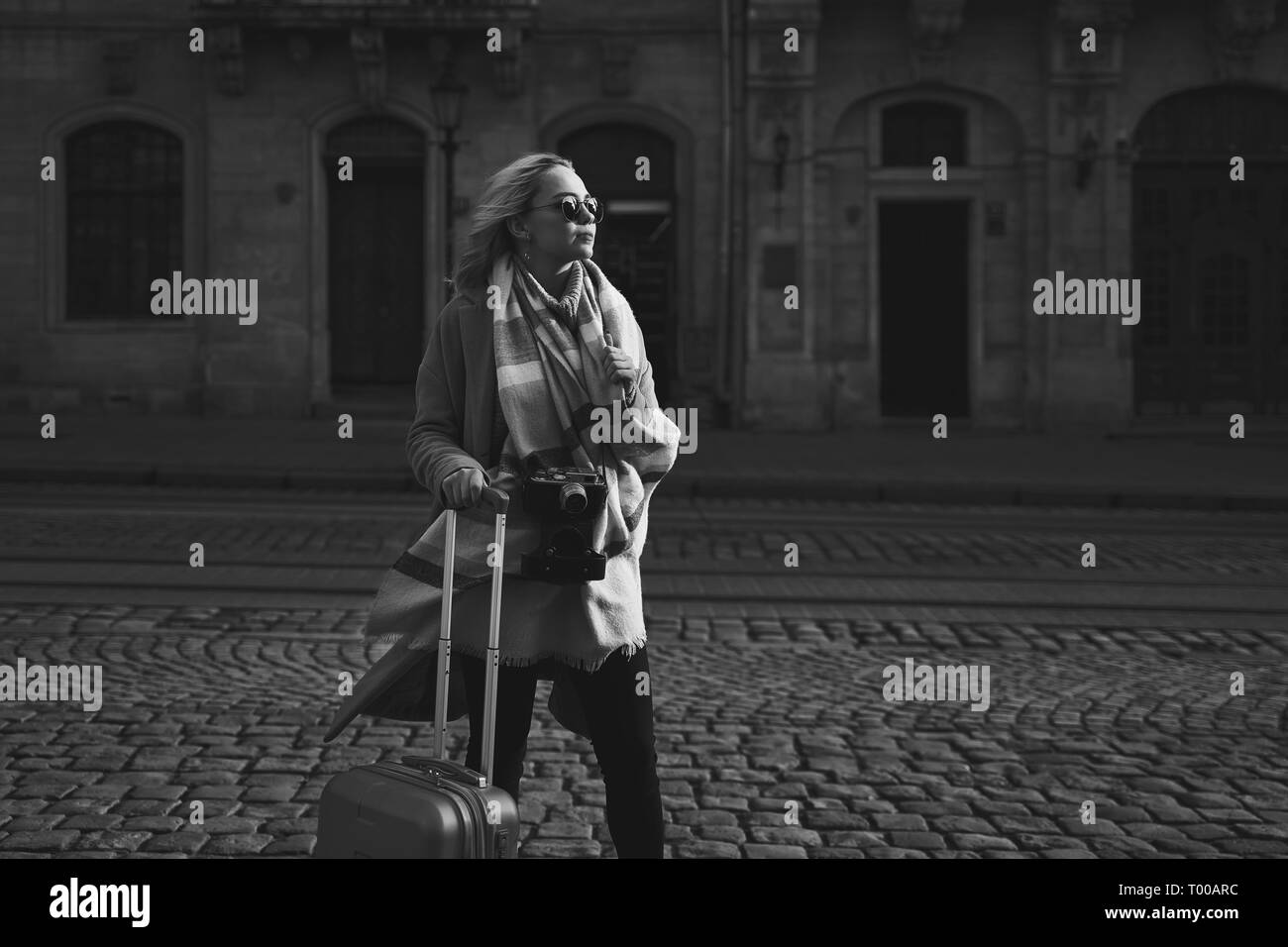 Young beautiful blond woman tourist with a wheeled travel bag and vintage film camera standing on Rynok square in Lviv, Ukraine Stock Photo