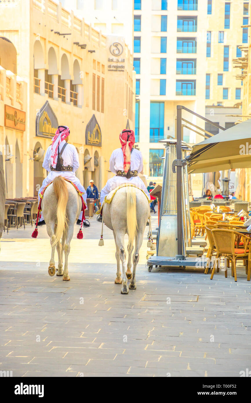 Doha, Qatar - February 20, 2019: two police on horse in traditional clothes at old Souq Waqif riding white Arabian Horses on pedestrian road. Popular Stock Photo