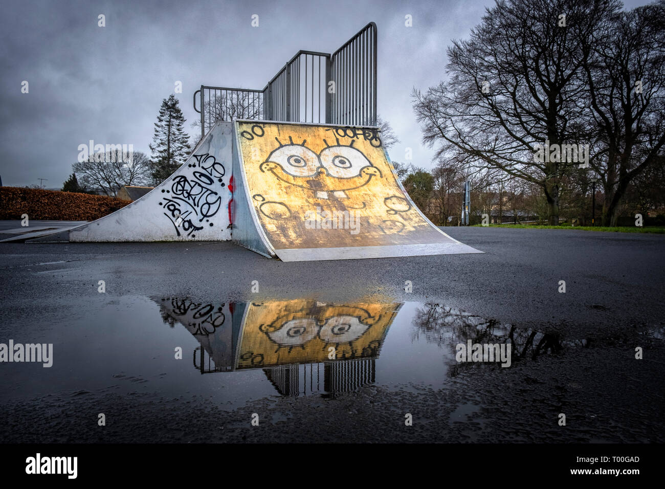 Empty children's playground with graffiti face painted on an deserted skateboard ramp reflected in puddle in dismal wet weather Stock Photo