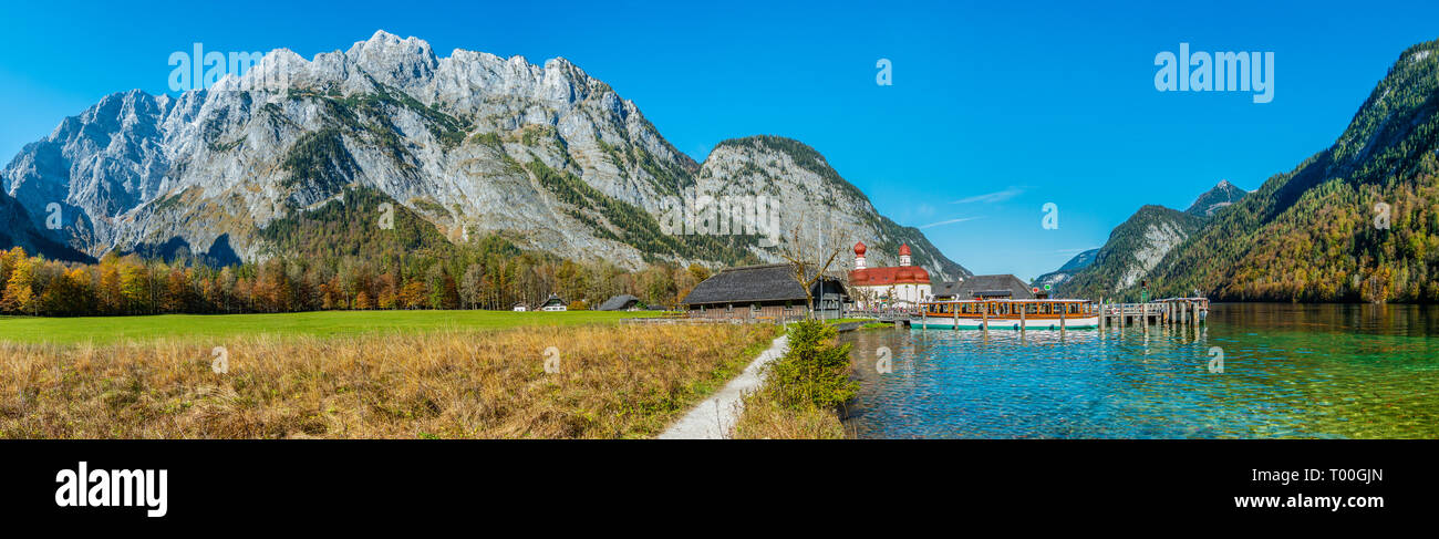 Panorama image of the Koenigssee with the chapel of st bartholomew and a tourist boat. In the background the illuminated mountain formation of Watzman Stock Photo